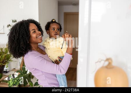 Lächelnde junge alleinerziehende Mutter, die Tochter trägt, die auf dem Kühlschrank in der Küche zu Hause schreibt Stockfoto