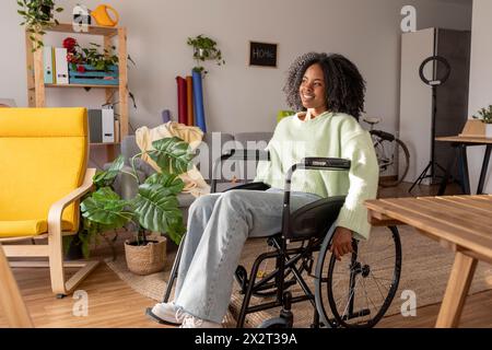 Lächelnde junge Frau mit Behinderung, die zu Hause im Rollstuhl sitzt Stockfoto