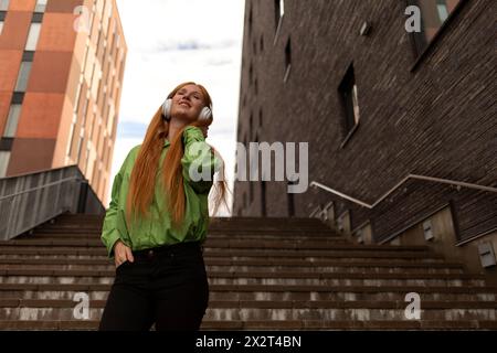 Lächelnde, schöne junge rothaarige Frau, die Musik hört, die auf Stufen steht Stockfoto