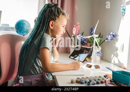 Mädchen mit gefärbten langen geflochtenen Haaren, die im Buch von Windturbinenmodellen am Tisch schreiben Stockfoto