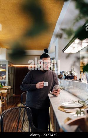 Kunde nutzt das Smartphone, das sich an der Bar mit einer Kaffeetasse im Restaurant lehnt Stockfoto
