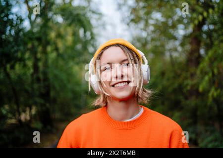 Lächelnde junge Frau, die kabellose Kopfhörer trägt und Musik im Park hört Stockfoto