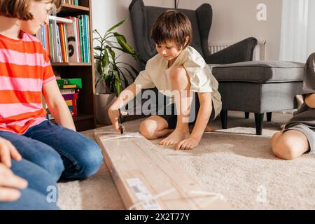 Boy Opening Plank Paket mit Schere von Geschwistern und Vater kniend im Wohnzimmer Stockfoto