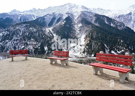 Zementierte Bänke mit roten Rückenlehnen mit Blick auf die Berge des Sangla Valley, Himachal Pradesh, Indien. Friedliche Außenlandschaft mit Schnee und Wald. Stockfoto