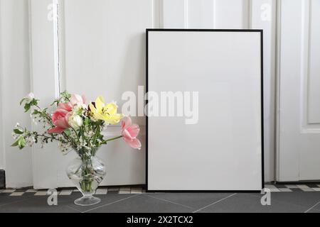 Stillleben im Frühling. Tulpen, Kirschblüten Blumenstrauß in Glasvase. Leerer schwarzer Bilderrahmen, Postermockup auf grauem Fliesenboden. Weiße Wand Stockfoto