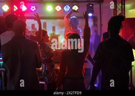 Clubber feiern und tanzen, während sie dj-Auftritte im Nachtclub besuchen. In der Diskothek stehen junge Menschen mit erhobenen Händen auf der Tanzfläche, beleuchtet mit Scheinwerfern Stockfoto