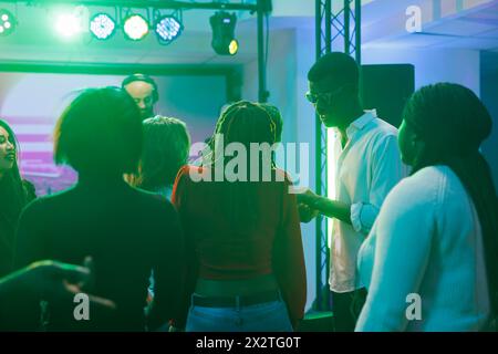 Leute, die feiern und Spaß bei geselligen Treffen haben, während sie ein dj-Konzert im Nachtclub besuchen. Vielfältige Leute entspannen sich auf der Tanzfläche, während der Musiker auf der Bühne im Dark Club auftritt Stockfoto