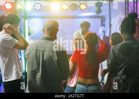 Junge Leute, die auf der Tanzfläche stehen, während sie ein Konzert mit elektronischer Musik im Nachtclub besuchen. Verschiedene Männer und Frauen tanzen während dj auf der Bühne mit farbenfrohen Spots Stockfoto