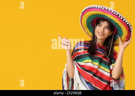 Junge Frau in Sombrero und farbenfrohem Poncho, die auf etwas auf gelbem Hintergrund zeigt. Cinco de Mayo-Feier Stockfoto