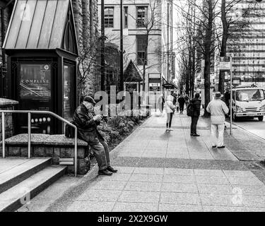 Vancouver, Kanada - 27. Februar 2024: Ein Schwarzweißfoto eines älteren Mannes in flacher Mütze, der auf sein Handy schaut, während er mit anderen an der Bushaltestelle wartete Stockfoto