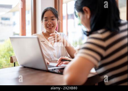 Zwei junge asiatische Studentinnen arbeiten zusammen an einem Projekt, schauen sich einen Laptop an und diskutieren, treffen sich in einem Café in Th Stockfoto