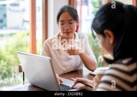 Zwei junge asiatische Studentinnen arbeiten zusammen an einem Projekt, schauen sich einen Laptop an und diskutieren, treffen sich in einem Café in Th Stockfoto