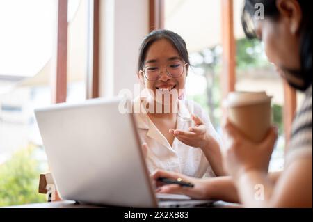 Zwei junge asiatische Studentinnen arbeiten zusammen an einem Projekt, schauen sich einen Laptop an und diskutieren, treffen sich in einem Café in Th Stockfoto