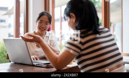 Zwei junge asiatische Studentinnen arbeiten zusammen an einem Projekt, schauen sich einen Laptop an und diskutieren, treffen sich in einem Café in Th Stockfoto