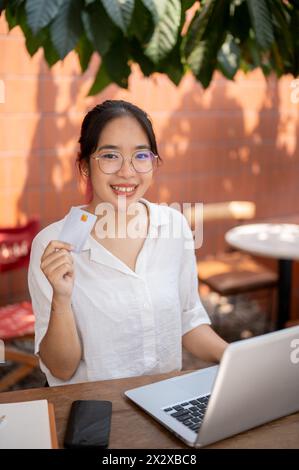 Eine lächelnde junge asiatische Frau in Freizeitkleidung zeigt ihre Kreditkarte vor die Kamera, während sie mit ihrem Computer an einem Café-Tisch sitzt und gehört Stockfoto
