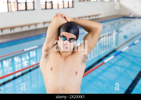 Kaukasischer junger männlicher Schwimmer, der sich vor dem Schwimmen in der Halle ausdehnt, Pool im Hintergrund Stockfoto