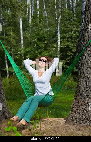 Die junge lächelnde Frau in dunkler Sonnenbrille sitzt in der Hängematte, wendet sein Gesicht zur Sonne und legt ihre Hände hinter ihren Kopf Stockfoto