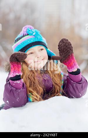 Ein lächelndes rosiges Mädchen in Jacke, Strickmütze und Fäustlingen liegt auf dem Schnee Stockfoto
