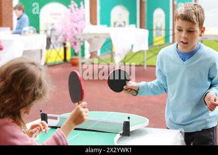 Kleines Mädchen und Junge in Blau spielen Tischtennis im Park am Sommertag. Konzentriere dich auf den Jungen. Stockfoto