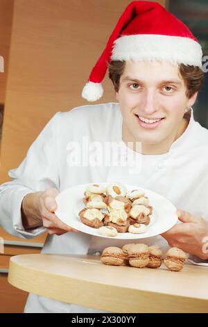 Ein junger lächelnder Konditor mit weihnachtsmann zeigt einen Teller mit Keksen und Kuchen darauf Stockfoto