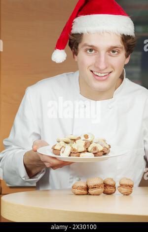 Ein lächelnder junger Konditor mit weihnachtsmann hält einen Teller mit Keksen und Kuchen Stockfoto