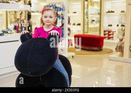 Das kleine Mädchen sitzt auf einem großen Plüschtier im Bekleidungsgeschäft Stockfoto