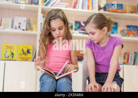 Zwei kleine Mädchen sind in der Bibliothek konzentriert und lesen ein Buch, konzentrieren sich auf das linke Mädchen Stockfoto