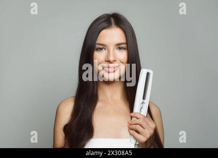 Nette junge gesunde Brünette Frau, die Haareisen hält und ihr langes glattes glänzendes Haar auf weißem Hintergrund glättet. Haarpflege, Friseur, Friseur Stockfoto