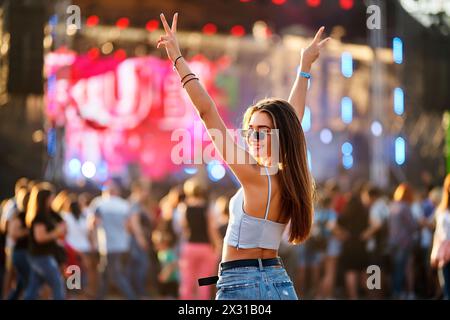 Glückliche junge Frau genießt Sommermusikfestival am Strand, Arme angehoben, Friedenszeichen. Die Menge tanzt in der Nähe der Bühne im Sonnenlicht. Partystimmung, Live-Konzert Stockfoto