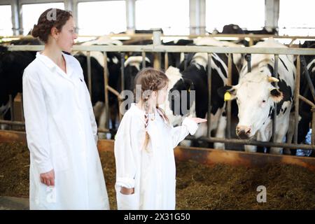 Mutter und Tochter im weißen Gewand reden im Stall mit vielen Kühen. Konzentriere dich auf Mädchen. Stockfoto