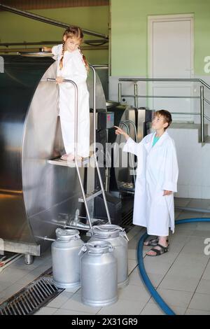 Ein lächelndes Mädchen und ein Junge in weißen Mänteln stehen neben einem großen Tank Milch in der Fabrik. Stockfoto