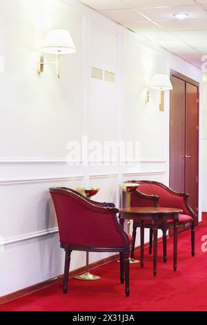 Zwei rote Sessel mit rundem Holztisch auf rotem Teppich im Flur des Hotels. Stockfoto