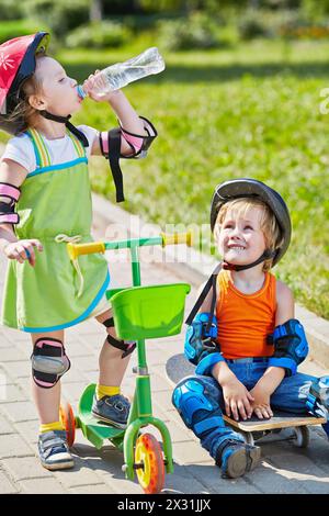 Der kleine Junge sitzt auf dem Skateboard und schaut das kleine Mädchen an, das Wasser aus einer Plastikflasche trinkt und mit einem dreirädrigen Roller steht Stockfoto