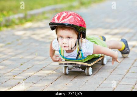 Das kleine Mädchen im Schutzhelm liegt auf dem Skateboard und blickt nach vorne, Arme entlang des Körpers. Stockfoto