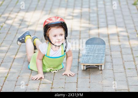 Das kleine Mädchen mit Schutzhelm liegt auf dem Parkweg in der Nähe des Skateboards und lehnt die Hände auf den Boden. Stockfoto