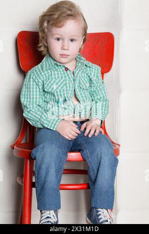 Ein kleiner Junge in einem karierten Hemd, der auf einem roten Stuhl an einer weißen Wand sitzt Stockfoto