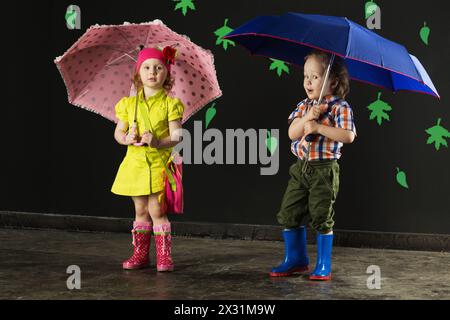 Kleiner Junge und Mädchen mit Regenschirmen in hellen Kleidern und Gummistiefeln Stockfoto