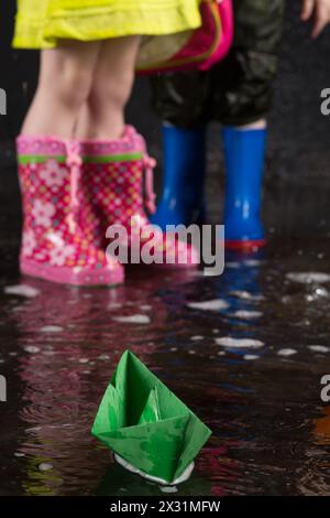 Grünpapierboot auf dem Hintergrund von Kinderfüßen in bunten Gummistiefeln Stockfoto