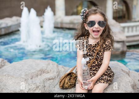 Kleines Mädchen in einem Leopardenkleid, das auf einem Felsen sitzt, mit einer Flasche neben dem Brunnen Stockfoto