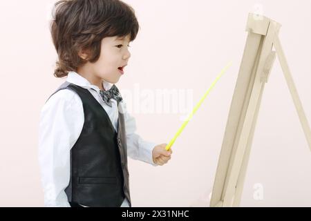 Der kleine brünette Junge mit gelbem Zeiger steht in der Nähe der Holzstaffelei im Studio. Stockfoto