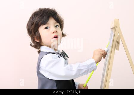 Kleiner Junge mit gelbem Zeiger zeichnet mit Kreide auf Tafel im Studio. Stockfoto