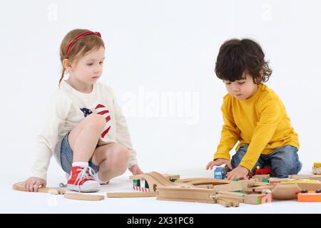 Kleiner Junge und Mädchen sitzen auf dem Boden und bauen Eisenbahn aus Holzteilen auf weißem Hintergrund. Stockfoto