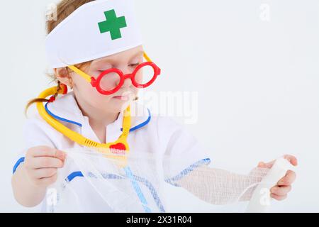 Ein kleines Mädchen, das als Krankenschwester verkleidet ist, spreizt Bandage auf weißem Hintergrund. Stockfoto