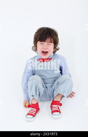 Kleiner Junge im blauen Jumpsuit sitzt auf dem Boden und weint auf weißem Hintergrund. Stockfoto