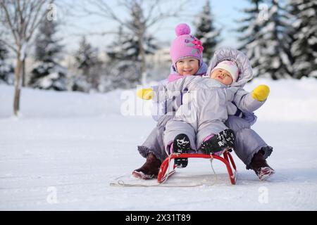 Kleines Mädchen und Baby in warmen Kleidern in der Nähe des Waldes sitzen im Winter auf rotem Schlitten. Stockfoto