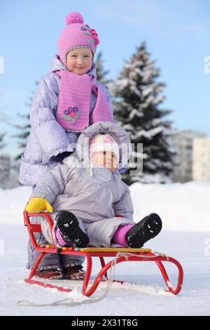 Das Baby in warmen Kleidern sitzt auf einem roten Schlitten und die kleine Schwester steht im Winter hinter ihm. Stockfoto