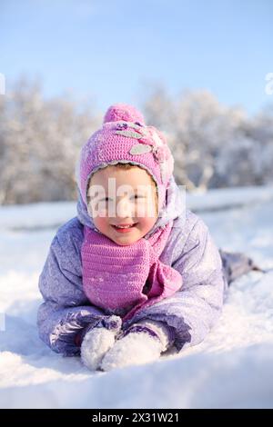 Kleines Mädchen in rosa Schal und Hut liegt auf dem Bauch auf Schnee und lächelt am Wintertag. Stockfoto