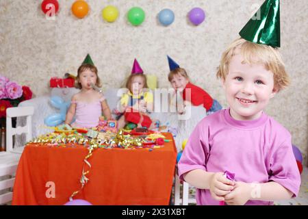 Junge in Mütze und mit Partybläsern lächelt auf der Geburtstagsfeier und drei Mädchen sitzen auf der Couch. Die Inschrift Happy Birthday (alles Gute zum Geburtstag) steht an der Wand. Konzentriere dich auf den Jungen. Stockfoto