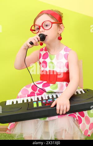 MOD-Mädchen in einem hellen Kleid und einer großen runden Brille, die in ein Mikrofon auf ihrem Schoßspielzeug-Piano singt Stockfoto