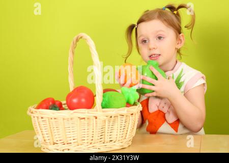Ein Mädchen mit Zöpfen, das mit Plastikgemüse spielt, das im Korb auf dem Tisch liegt Stockfoto